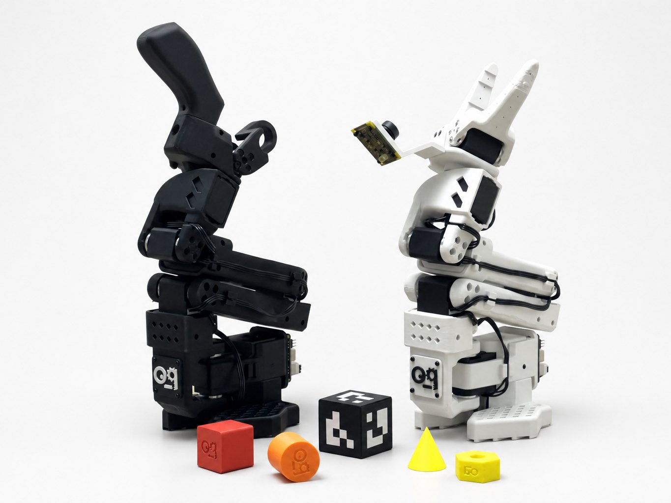 The SO-ARM101 Black & White pair: the black leader arm and the white follower arm with a camera, with the training objects and the ArUco cube in front of them